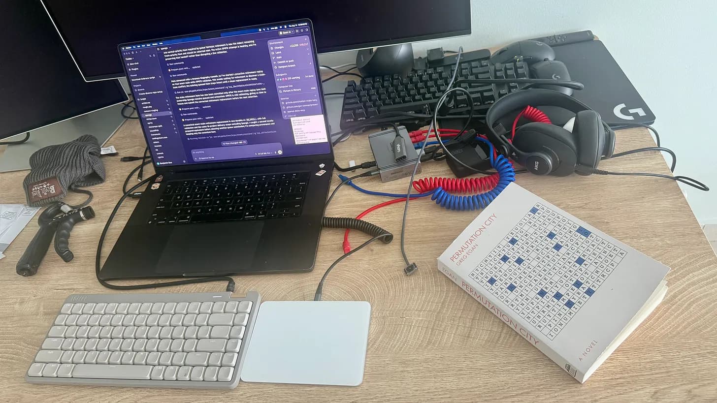 A laptop running coding agents on a desk beside a copy of Permutation City.