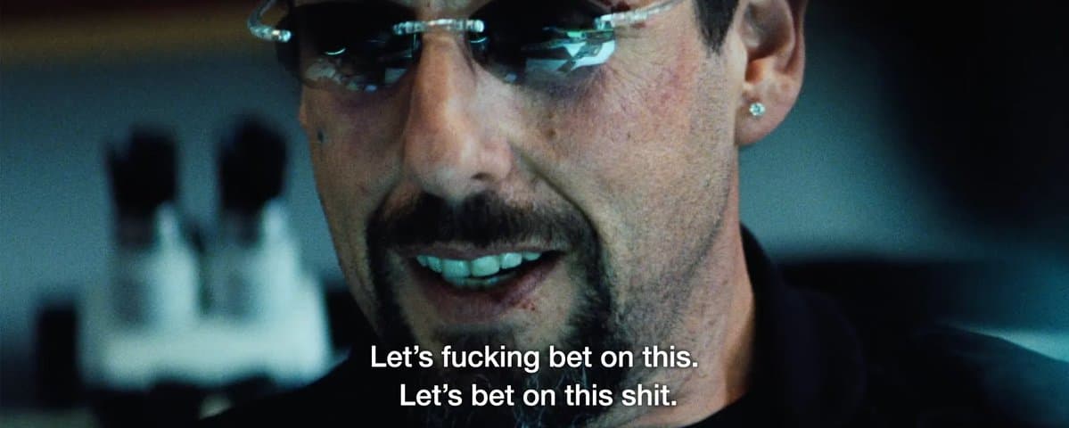 Close-up of Adam Sandler as Howard Ratner in Uncut Gems, grinning intensely in rimless glasses with a diamond stud earring. White subtitles read Let's fucking bet on this. Let's bet on this shit.