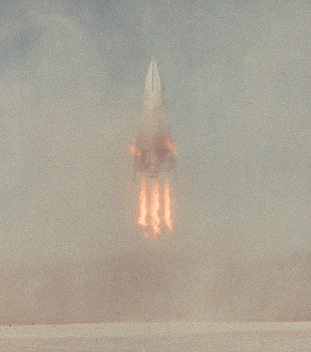 Grainy photograph of the DC-X Delta Clipper on four engine plumes through haze above a pale horizon.